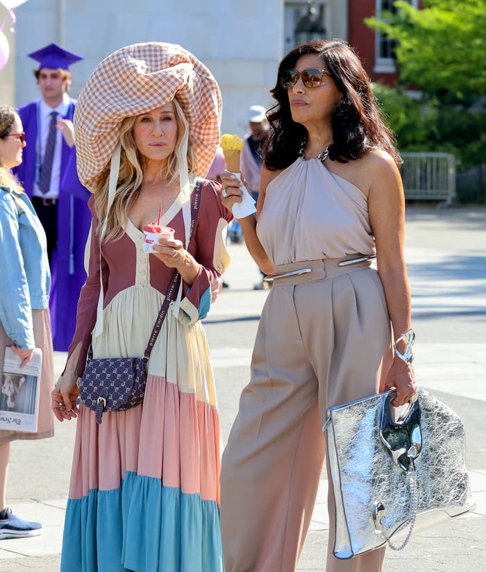 Sarah Jessica Parker and Sarita Choudhury holding ice cream cones, walking outdoors. Sarah wears a patchwork dress and large hat, Sarita wears a sleeveless jumpsuit