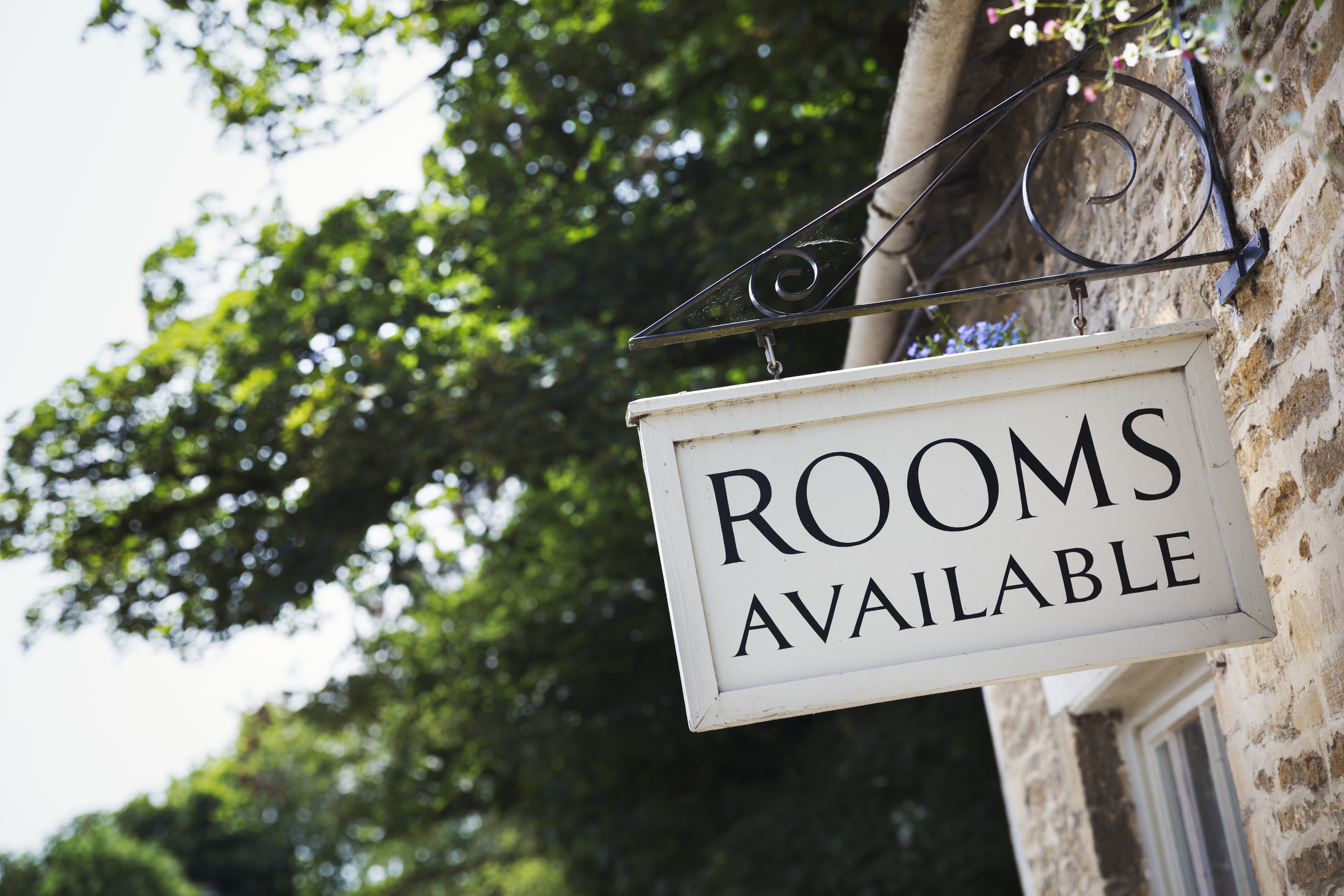 A sign hanging outside of a building reads "Rooms Available." The background shows trees and part of the building