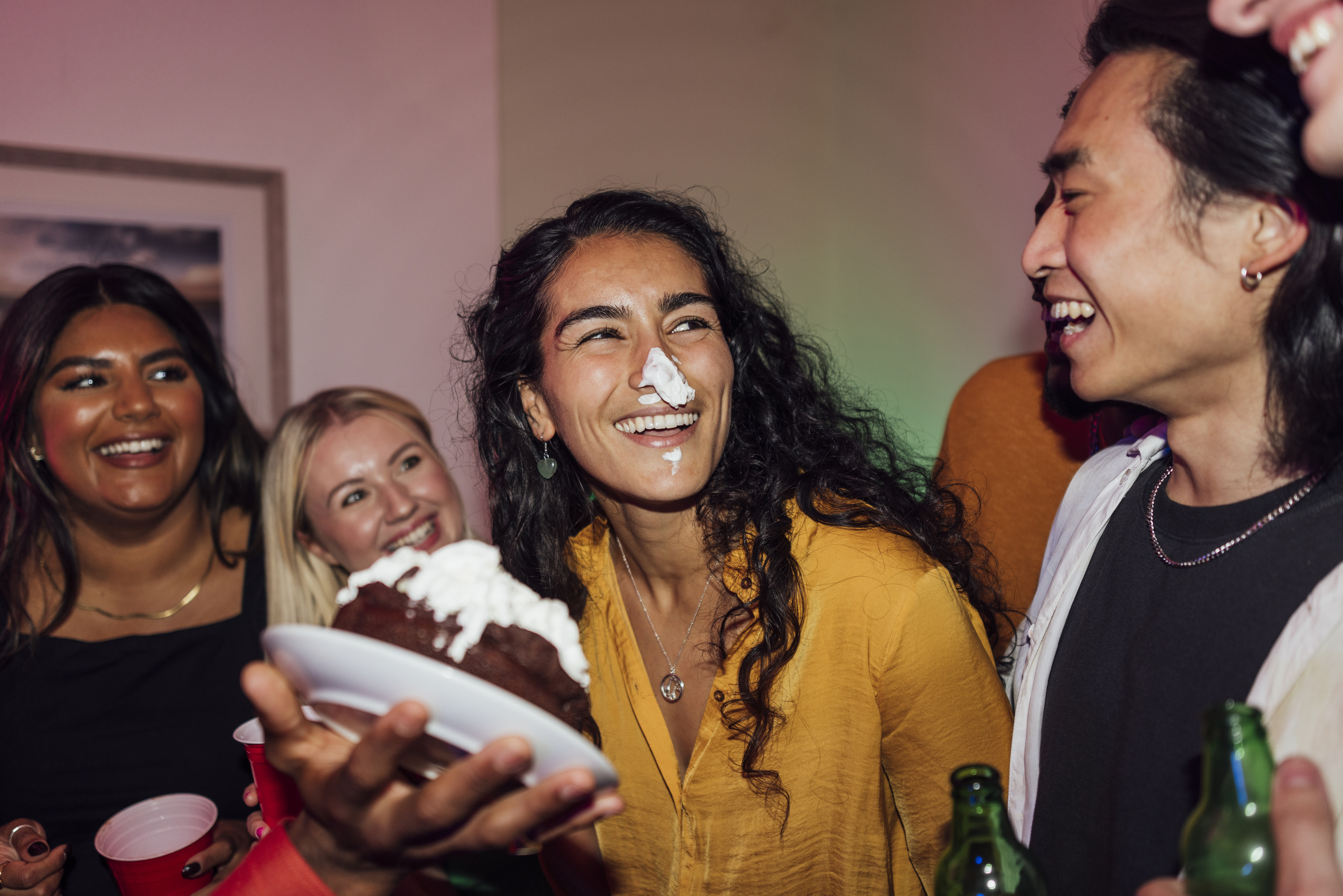 Group of friends at a party, one with pie on their face, smiling and holding a pie slice while others laugh. Names not given