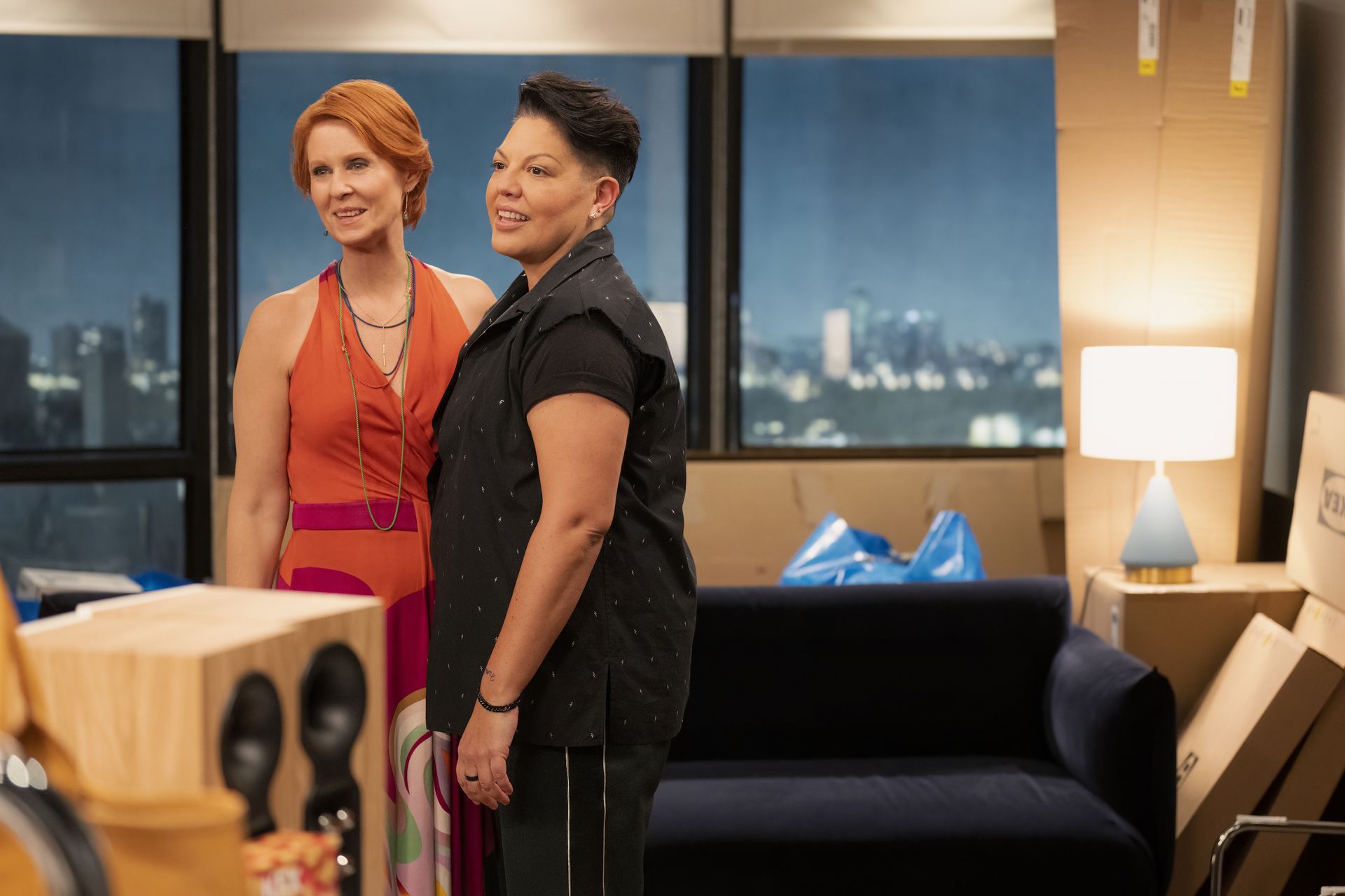 Cynthia Nixon in a dress and Sara Ramirez in a shirt and pants are standing together in an apartment with unpacked boxes