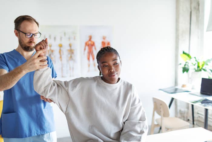 A doctor assists a patient by raising her arm in a medical office. Anatomical posters are visible in the background