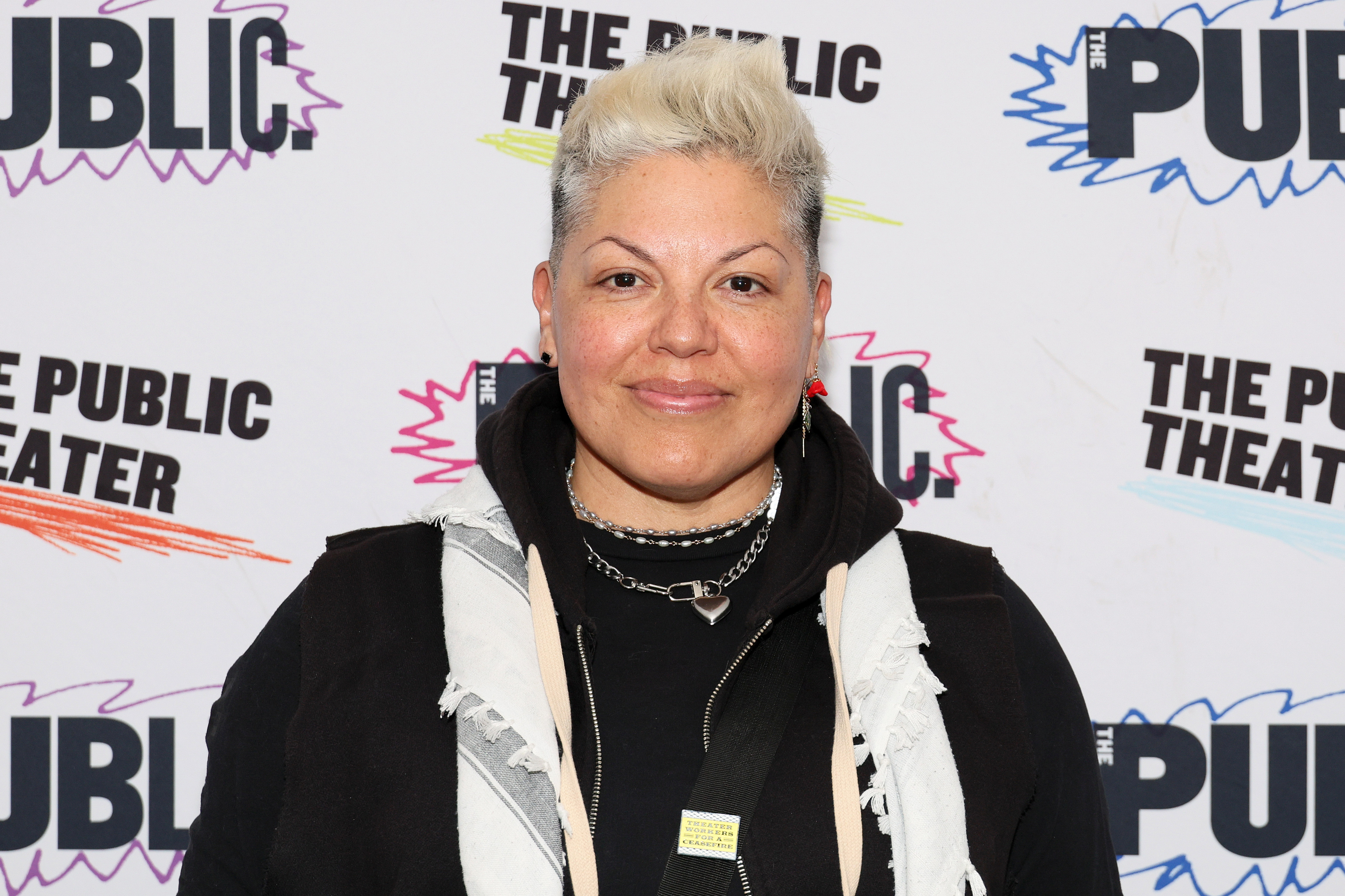 Sara Ramírez at The Public Theater event, wearing a hoodie with layered accessories including necklaces and a scarf