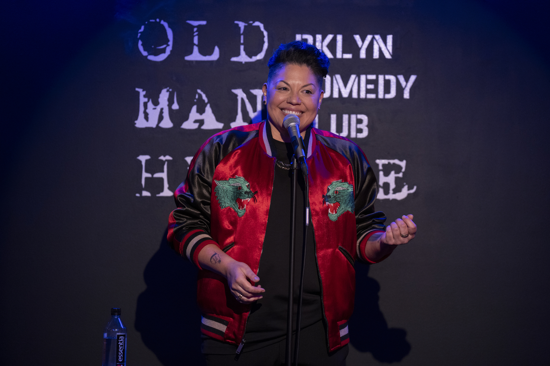 A performer wearing a jacket speaks into a microphone on stage at the Old Man Hustle BKLYN Comedy Club