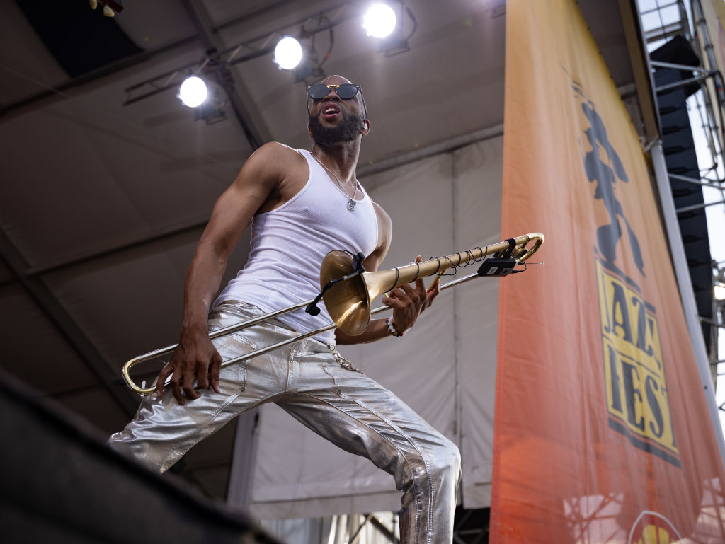 Man in a white tank top and metallic pants plays a trombone on stage at a jazz festival