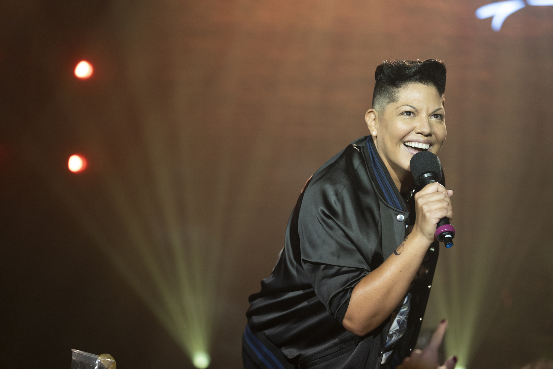 Sara Ramirez smiles while holding a microphone on stage during a performance