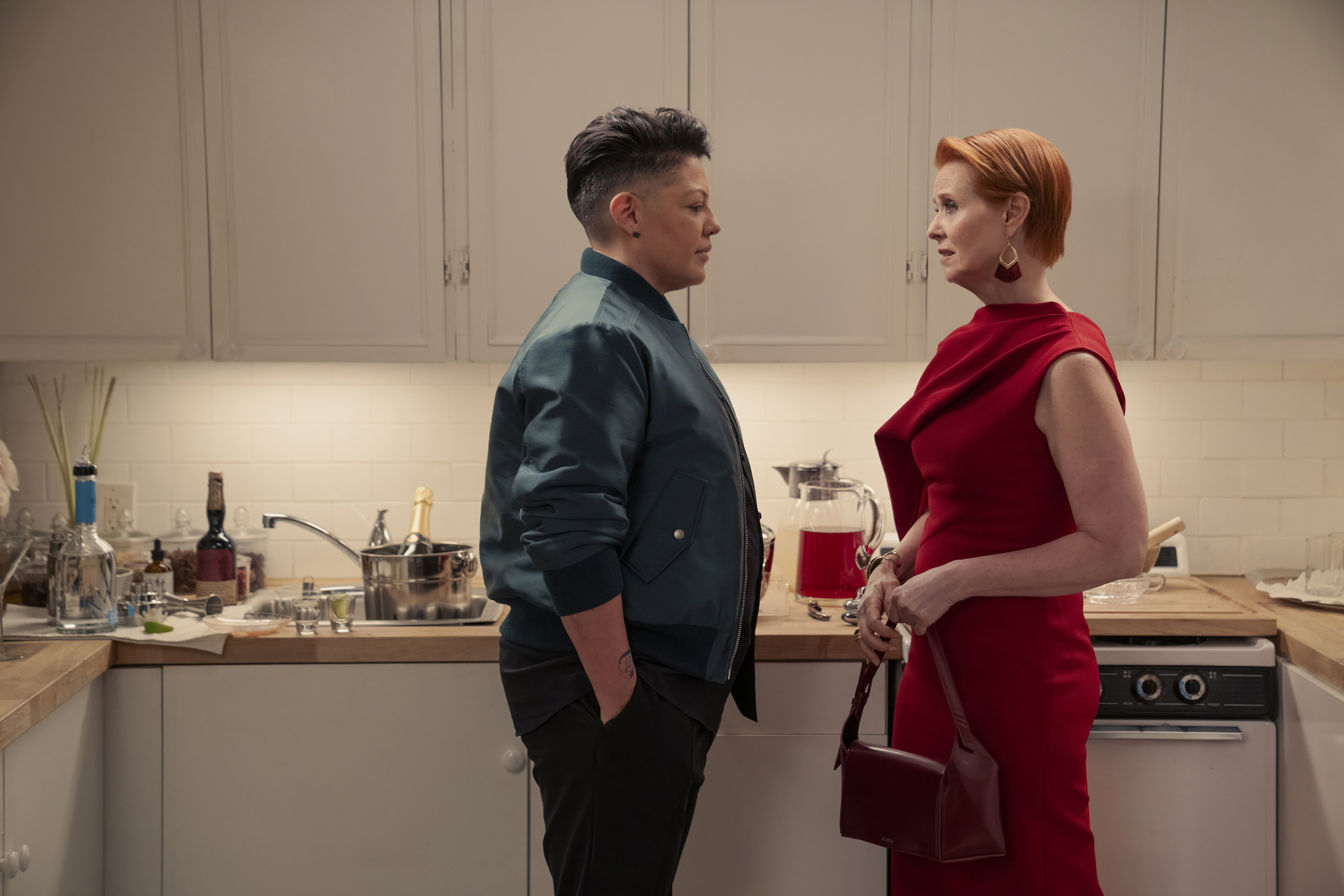 Sara Ramirez in a green jacket and Cynthia Nixon in a red dress stand in a kitchen, engaged in conversation