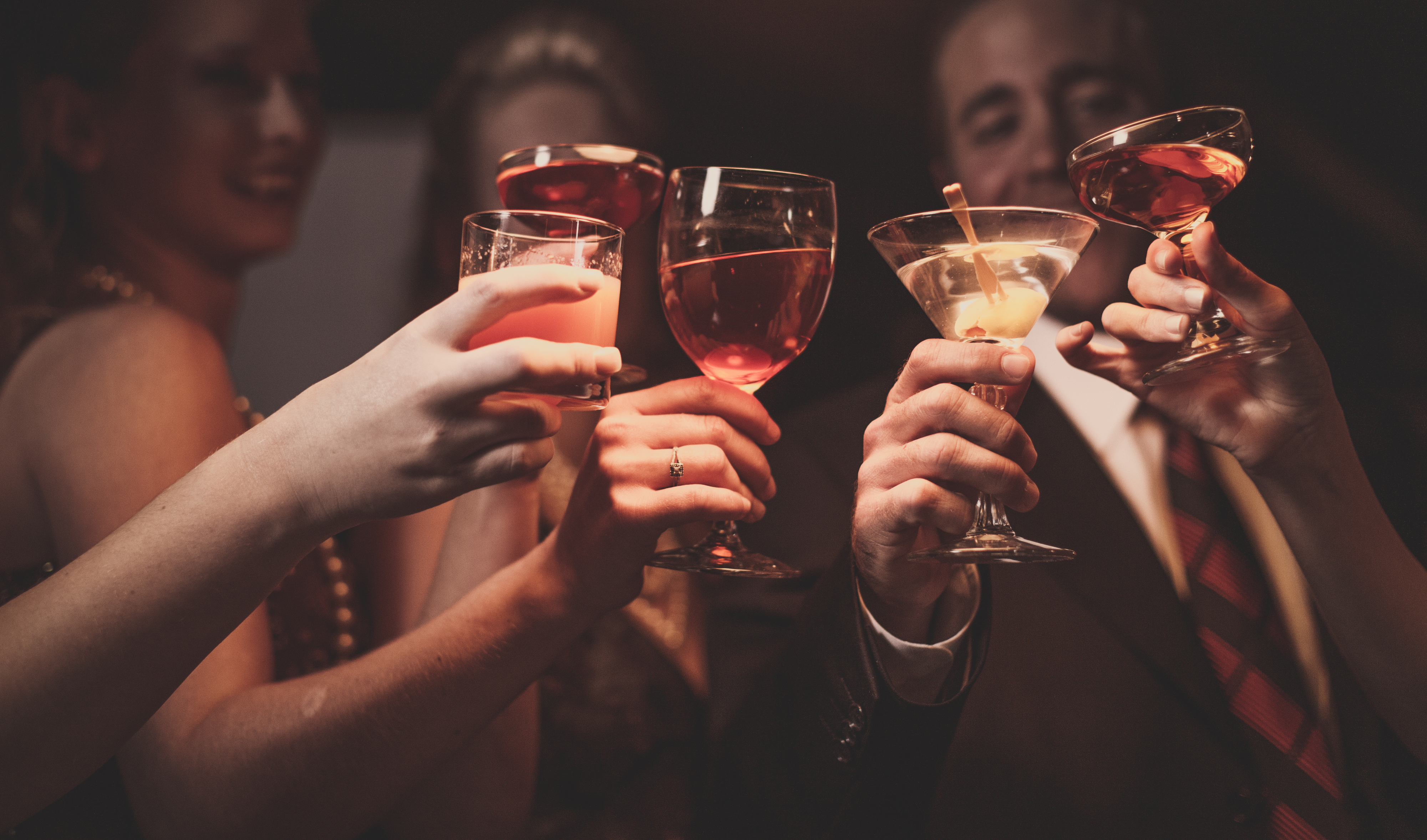 Several people clink glasses filled with various drinks during a social gathering. Their faces are blurred or not fully visible