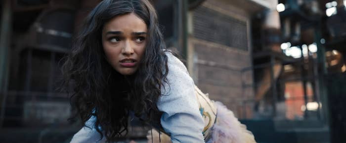 Rachel Zegler is in a distressed pose on the ground wearing a light outfit with a tulle skirt in an industrial area