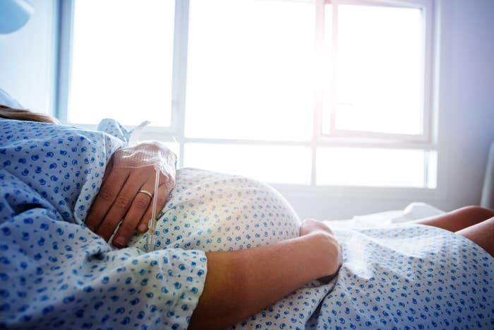 Close-up of a pregnant woman in a hospital gown lying on a bed, with sunlight streaming through a window