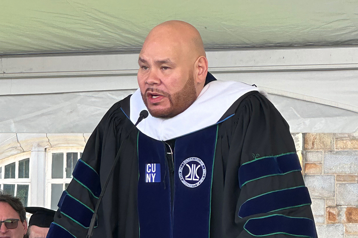 Fat Joe delivering a commencement speech at a graduation ceremony, wearing a traditional academic gown and hood on stage
