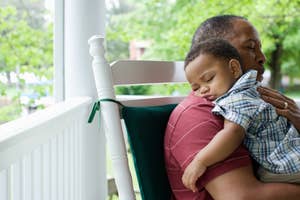 A man sits on a porch rocking chair, holding a young child who is sleeping on his shoulder. The setting is peaceful with a green, leafy background