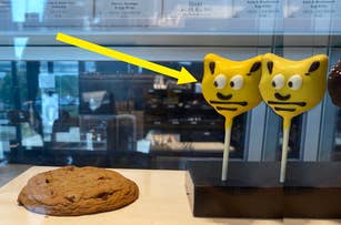 Two yellow cat-shaped cake pops with expressive faces on sticks next to a chocolate chip cookie in a display case