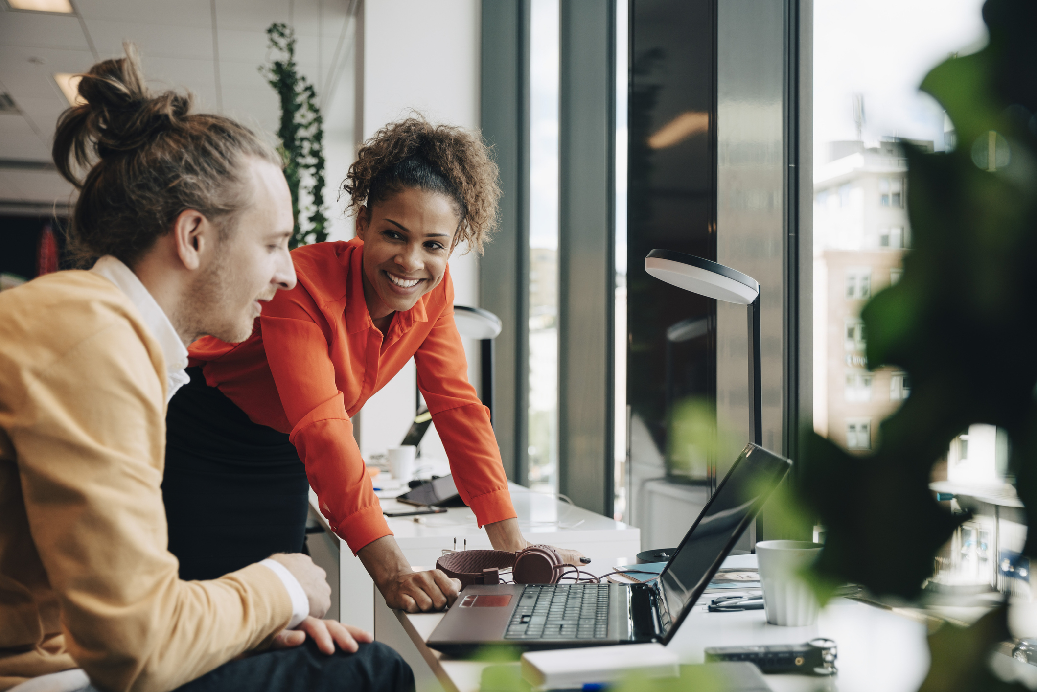 A man with a bun and a woman with curly hair are smiling and working together at a desk with a laptop and headphones near a window in an office setting