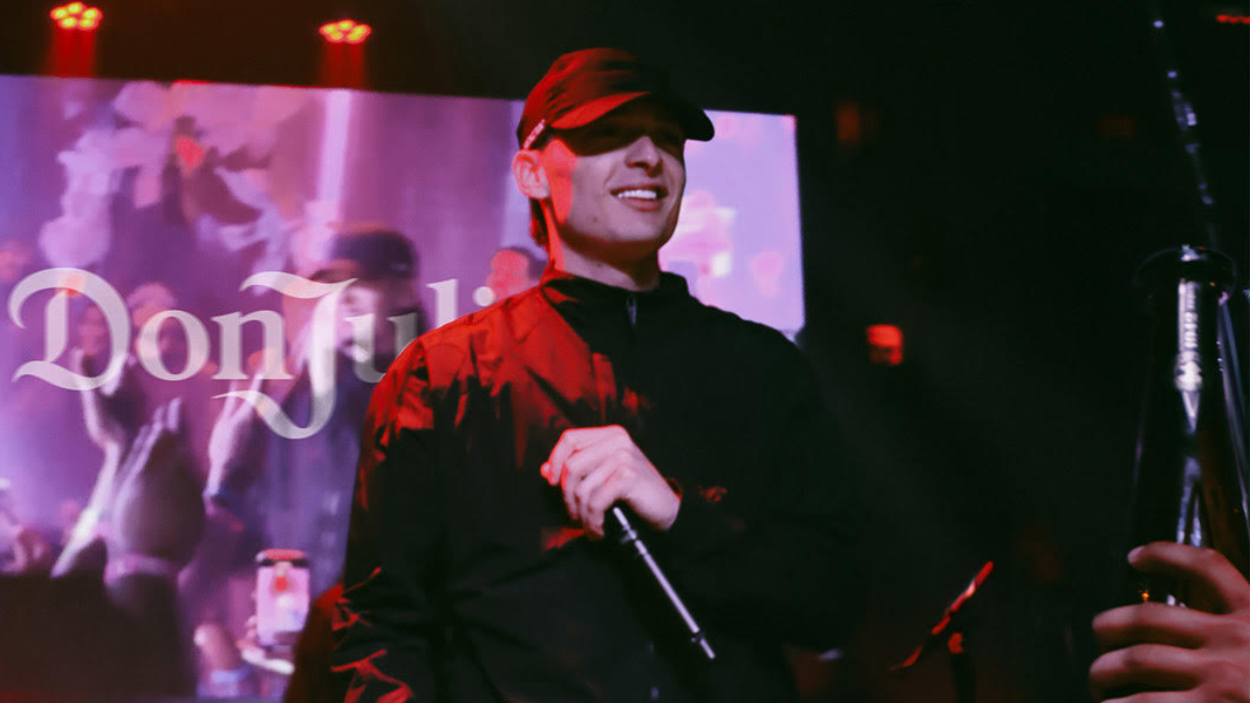 Peso Pluma in a black jacket and black cap performs on stage with a microphone. Signage in the background reads &quot;Don Julio&quot;