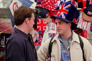 A man wearing a backpack and a large Union Jack hat talks to another man in front of a street vendor selling souvenirs with British flags