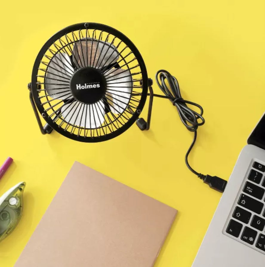 Kipas Holmes Desk Black Holmes dicolokkan ke laptop, di samping buku catatan, dispenser pita, dan pena di latar belakang kuning