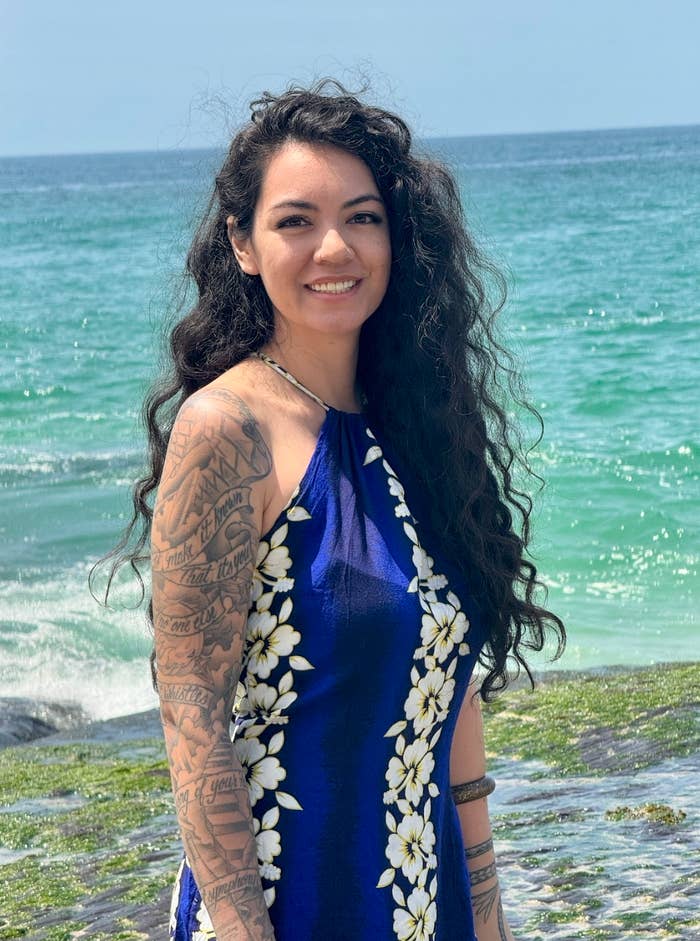 Woman with long curly hair, tattooed arm, wearing a floral halter dress, smiles while standing by the ocean