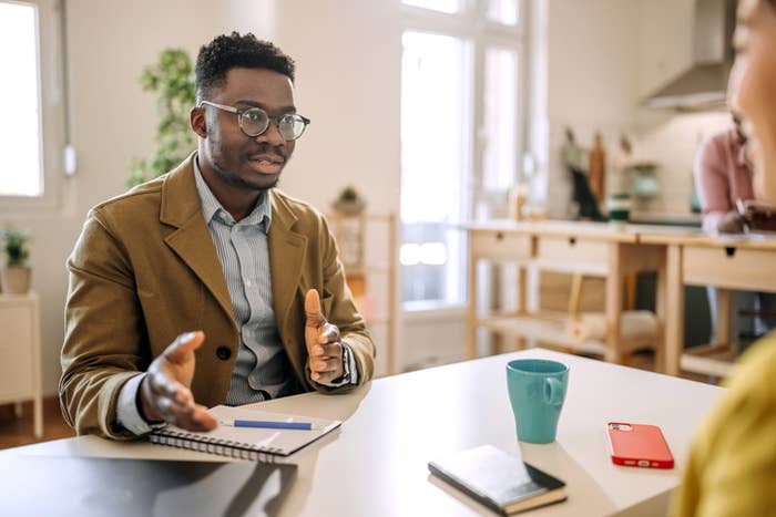 A person wearing glasses and a suit jacket sits at a table, gesturing while speaking to another person in a casual top. There are notebooks and a coffee cup on the table