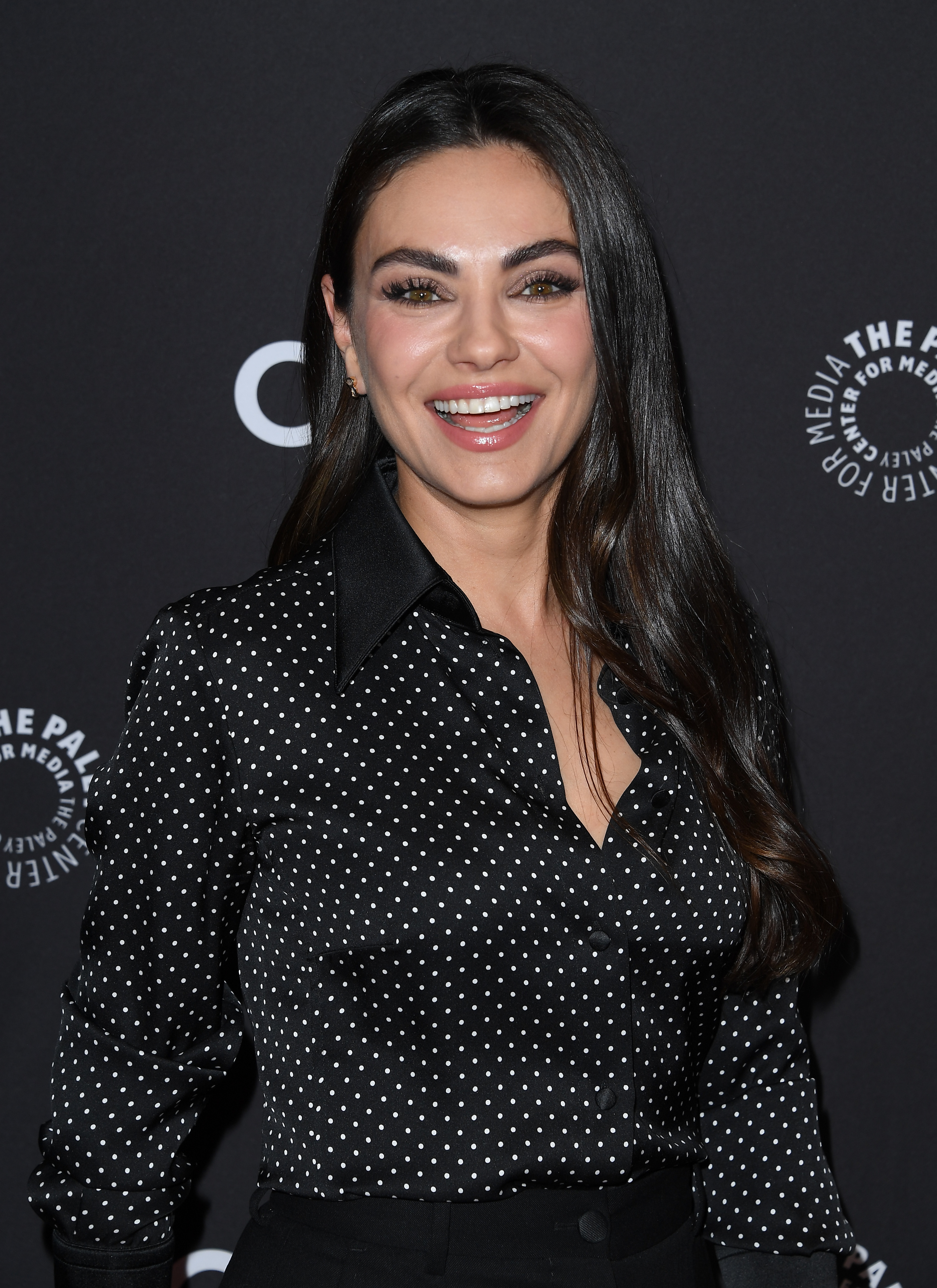 Mila Kunis smiles for the camera, wearing a stylish polka-dot blouse, at a media event