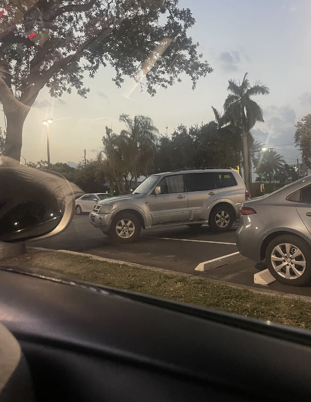 A parked SUV and a car viewed from inside another vehicle in a parking lot with trees and streetlights. No people are visible