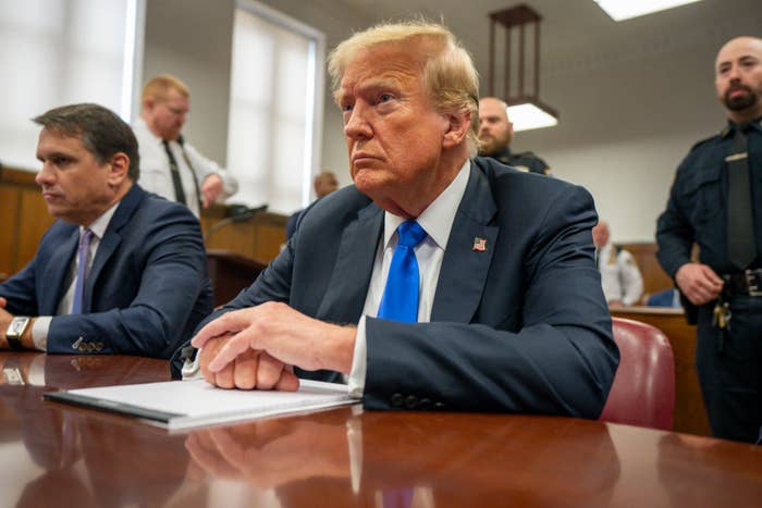 Donald Trump sits at a courtroom table with a notepad, wearing a suit and blue tie. Others are present, including his lawyer seated beside him