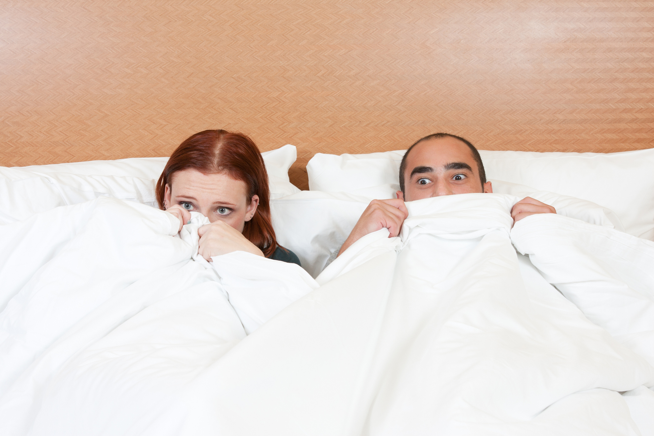 Two people in bed, partially hiding under white covers with surprised expressions, looking towards the camera