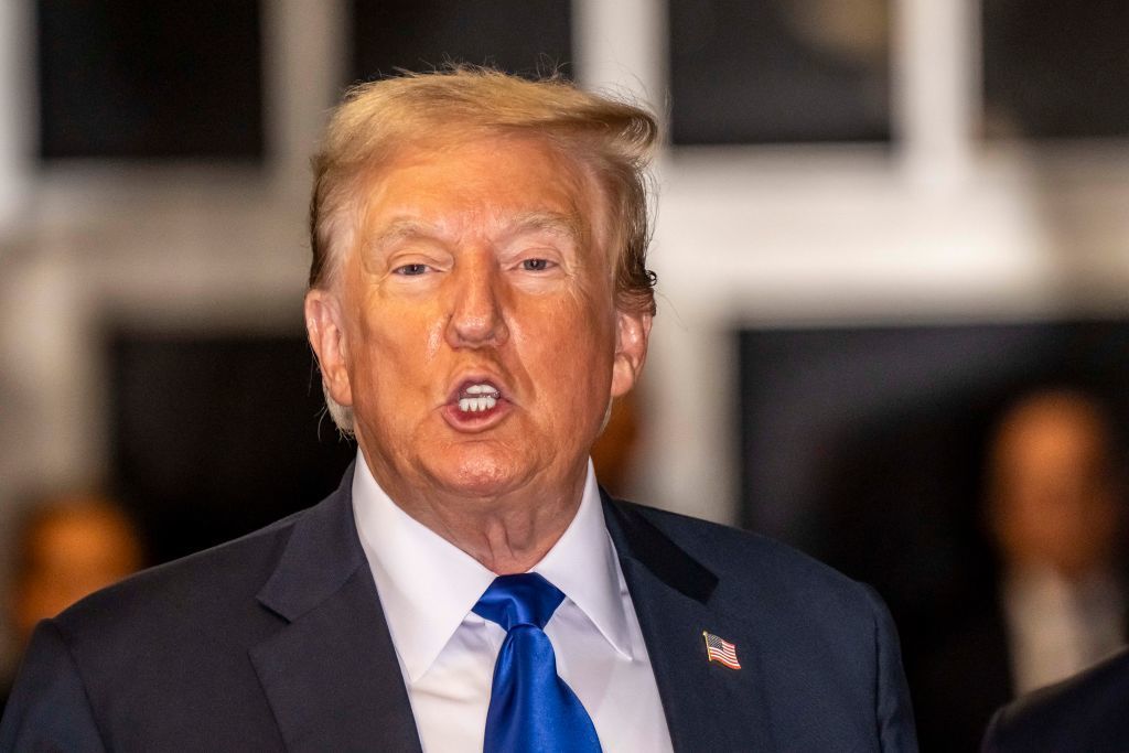 Donald Trump wears a suit and blue tie, speaking with an intense expression in a public setting