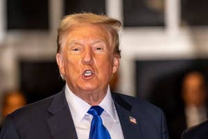 Donald Trump wears a suit and blue tie, speaking with an intense expression in a public setting