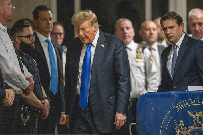 Donald Trump stands surrounded by men in suits and security personnel in a formal setting