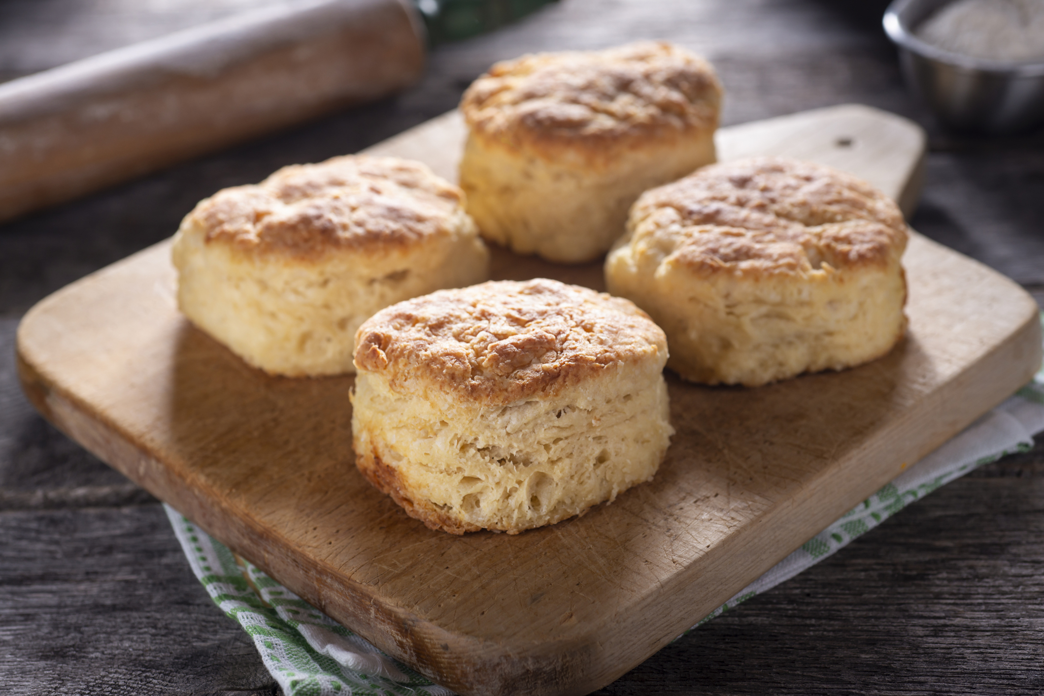 Four freshly baked biscuits are on a wooden cutting board with a rolling pin in the background