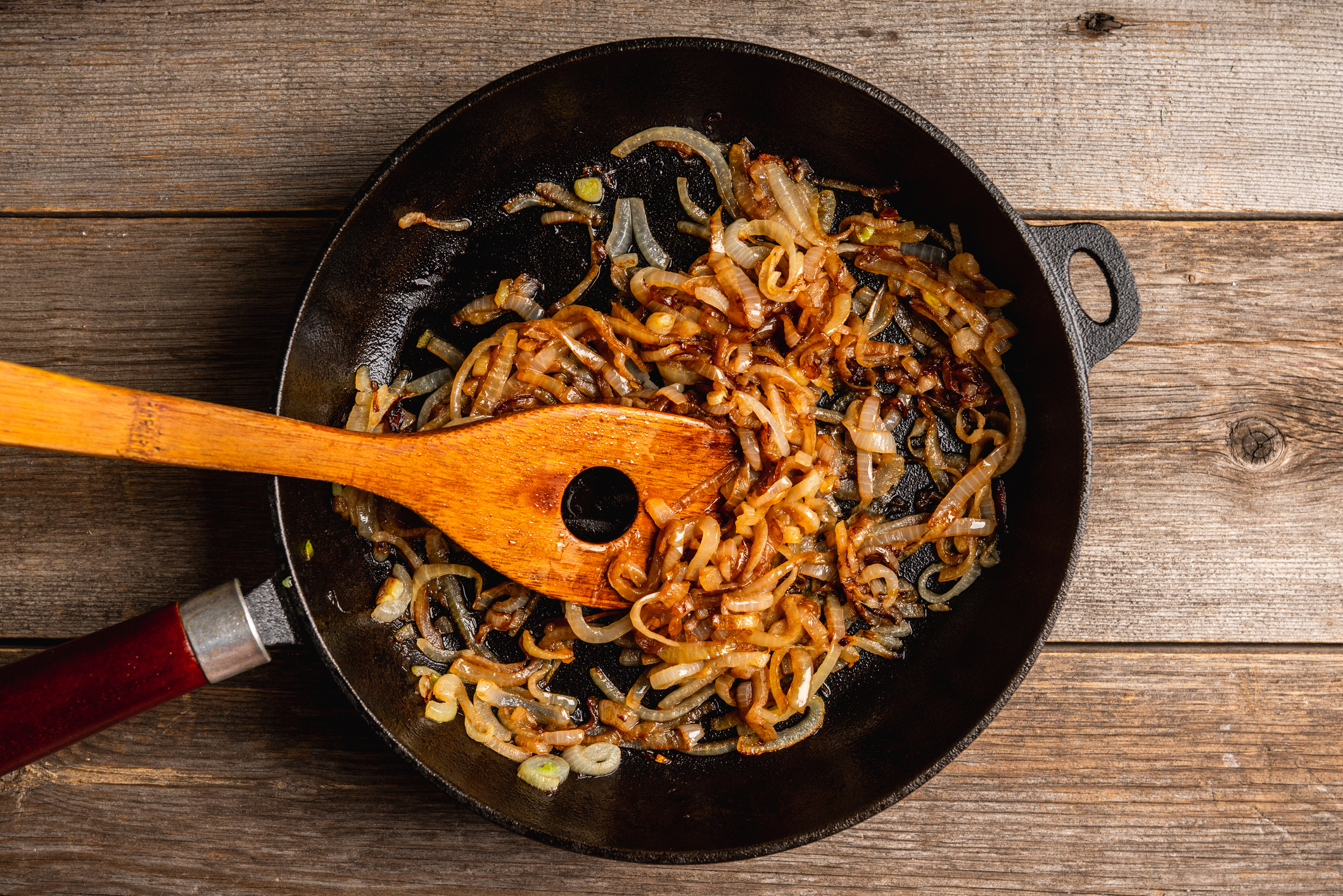 A wooden spoon stirring caramelized onions in a cast iron pan on a wooden table
