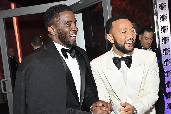 Sean Combs and John Legend are smiling and shaking hands at a formal event. Sean is wearing a black suit with a cape, and John is in a white tuxedo