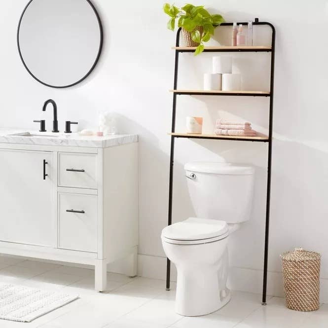 Bathroom with over-the-toilet storage shelf holding towels, plants, and toiletries. A white vanity with a round mirror and a black faucet is beside the toilet