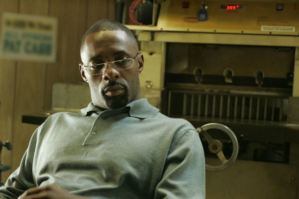 A man sits thoughtfully in an industrial setting, wearing glasses and a grey polo shirt