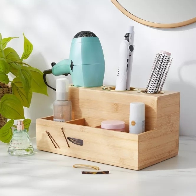 Bamboo organizer holds a turquoise hair dryer, white curling iron, round brush, facial mist, cream, and hair accessories on a bathroom counter