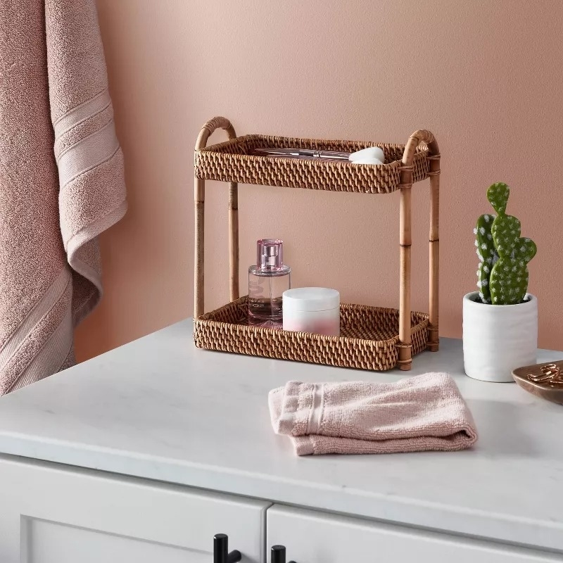A small rattan shelf on a bathroom counter holding a bottle of perfume, a candle, and a small sponge, next to a potted cactus, a towel, and a bowl
