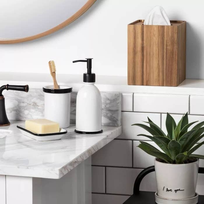 Modern bathroom countertop with a black pump soap dispenser, toothbrush holder, bar of soap, wooden tissue box, and a potted plant