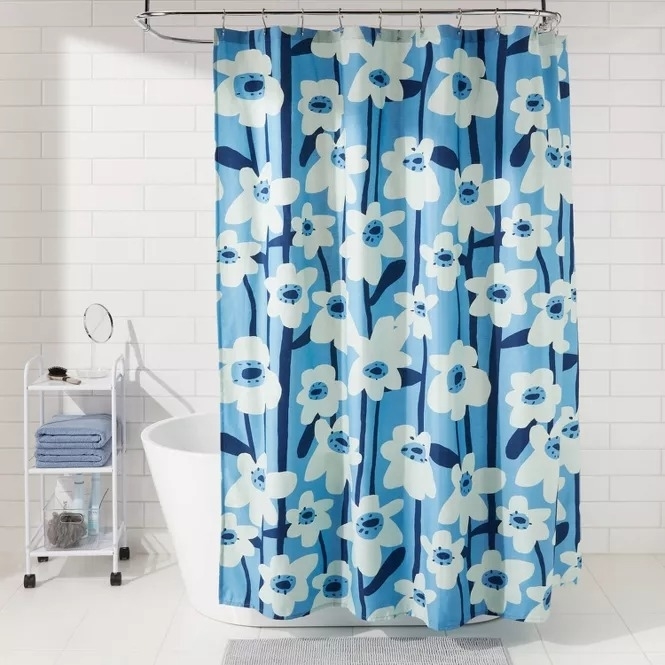 Shower curtain with large floral print in various shades of blue hangs in a modern white-tiled bathroom. Accessories on a nearby shelf include towels and toiletries