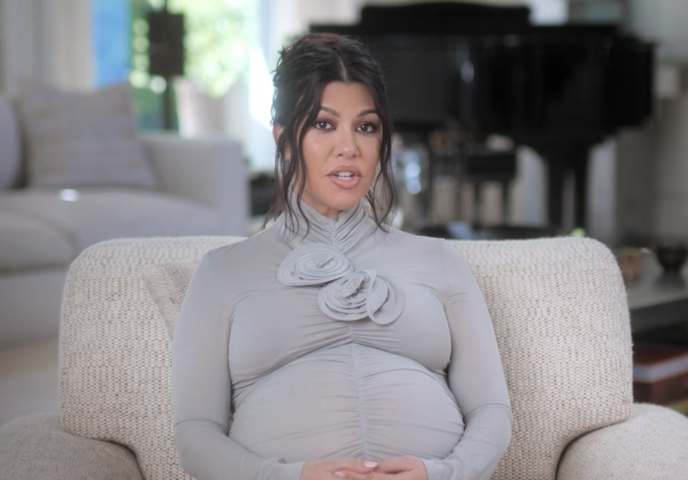 Kourtney Kardashian sits on a beige couch in a modern living room, wearing a form-fitting light turtleneck dress with decorative flower details on the chest