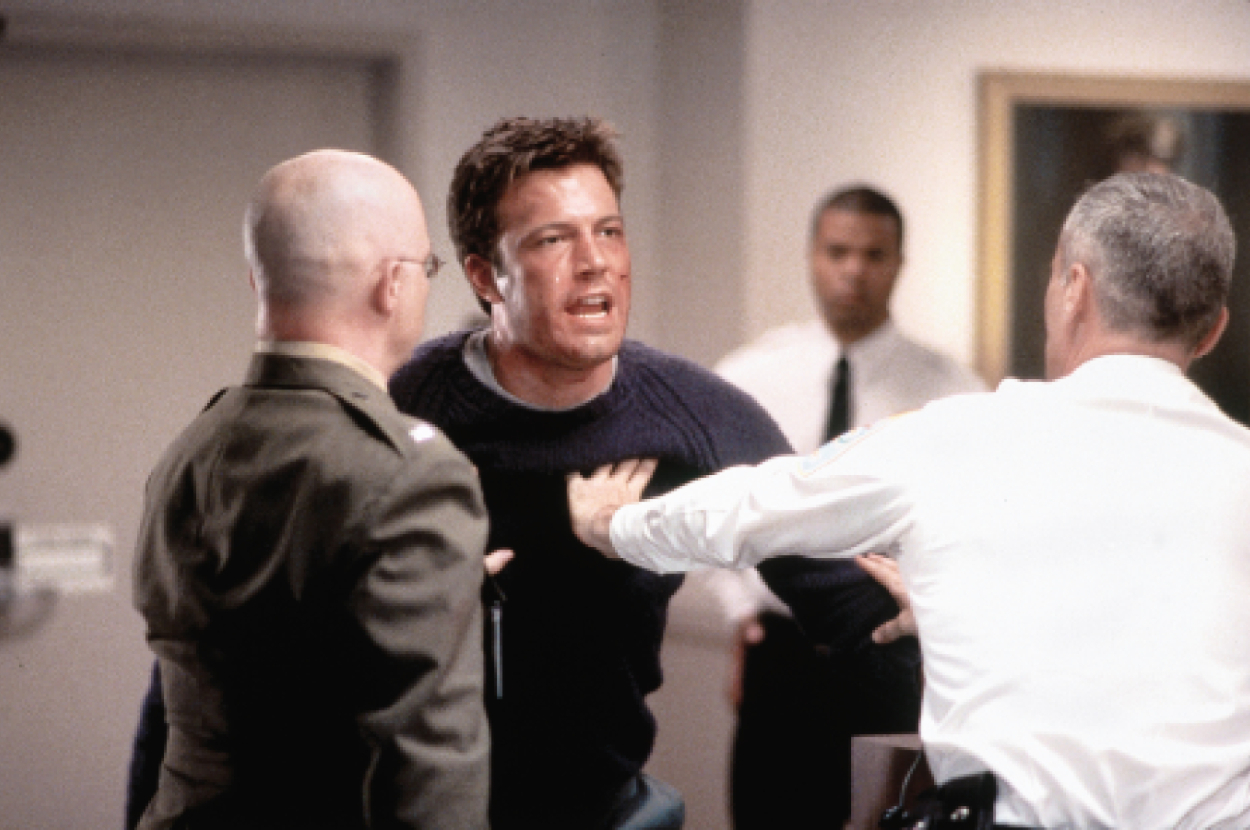 Ben Affleck, restrained by two uniformed men, looks emotional in a tense indoor setting. A man in the background observes