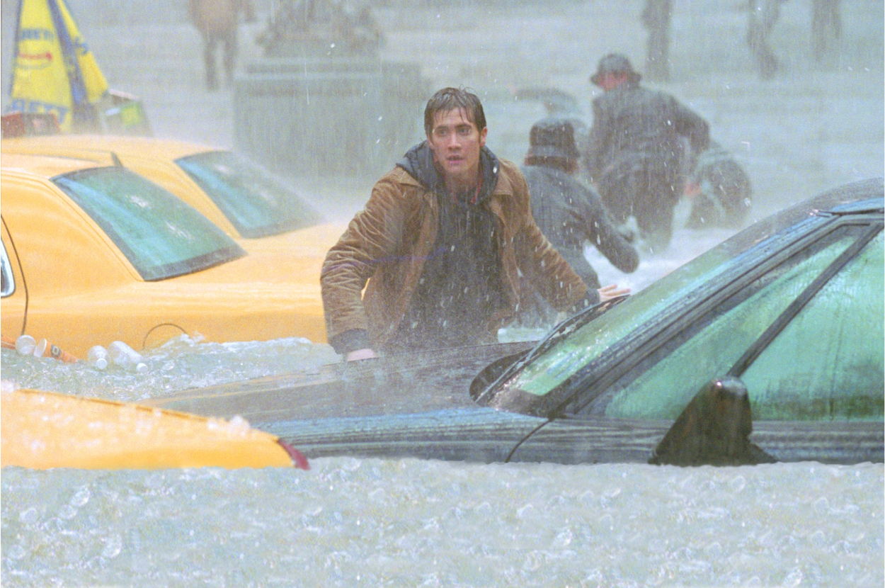 A man, wearing a brown jacket and looking distressed, wades through floodwaters between cars in a city scene during heavy rainfall