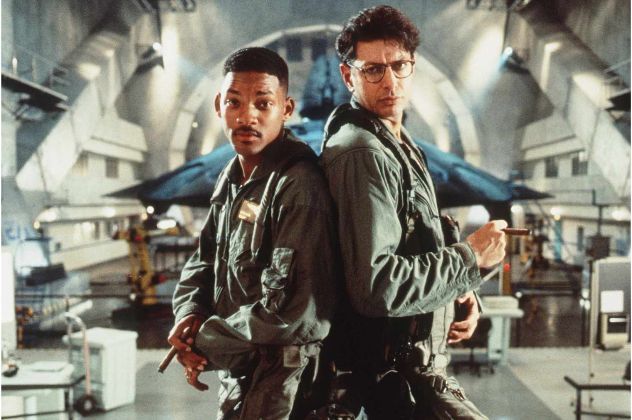 Will Smith and Jeff Goldblum stand back to back in a futuristic setting, each holding a cigar and wearing military-style jackets