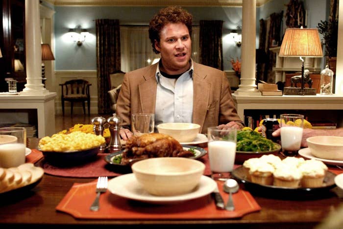 Seth Rogen, dressed in a casual suit, sits at a dining table filled with various foods, including chicken, macaroni, and cupcakes, looking surprised
