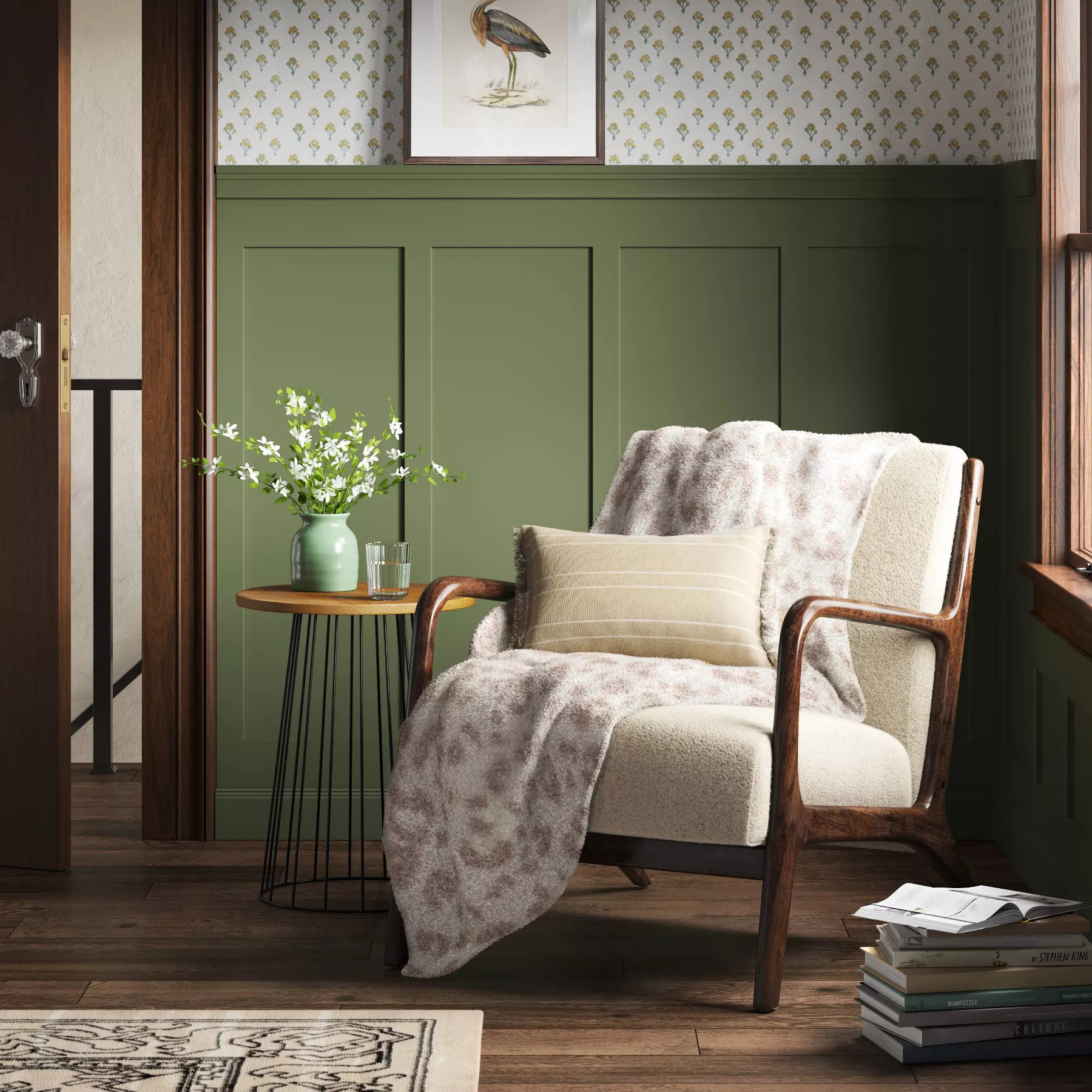 Cozy reading nook featuring the throw blanket over an armchair next to a side table with flowers, and framed artwork on the wall. Green wainscoting and wood accents