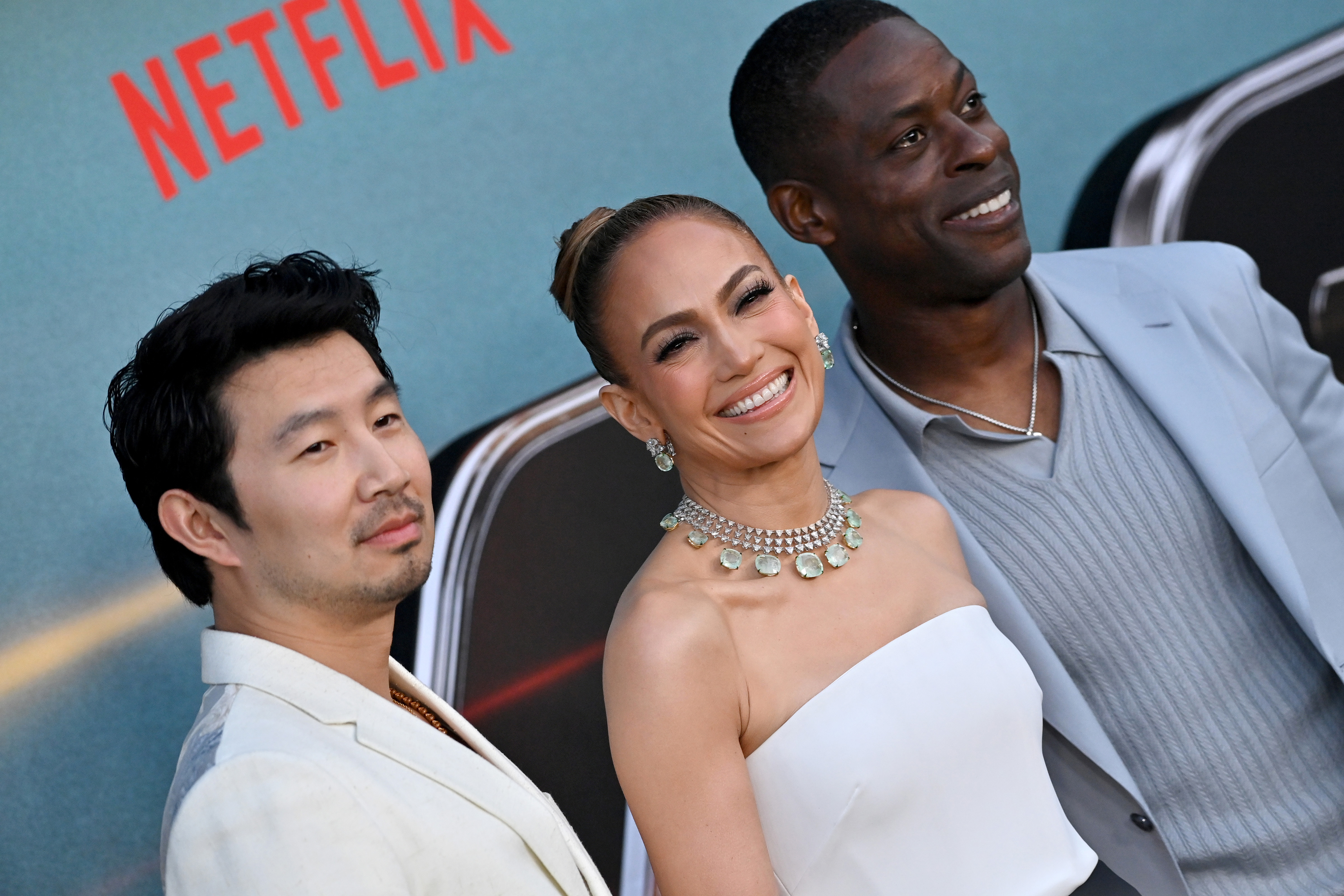Simu Liu, Jennifer Lopez, and Sterling K. Brown pose at a Netflix event, with Jennifer wearing an elegant strapless white dress and a statement necklace