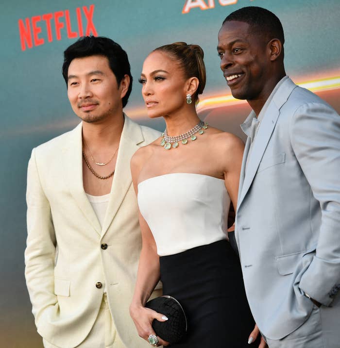 Simu Liu, Jennifer Lopez, and Sterling K. Brown pose on the red carpet. Jennifer wears an elegant strapless dress with a necklace, Simu and Sterling wear stylish suits