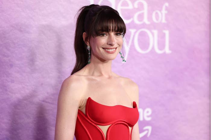 Anne Hathaway stands on the red carpet in a strapless, form-fitting gown with a unique neckline, smiling for the cameras at an event