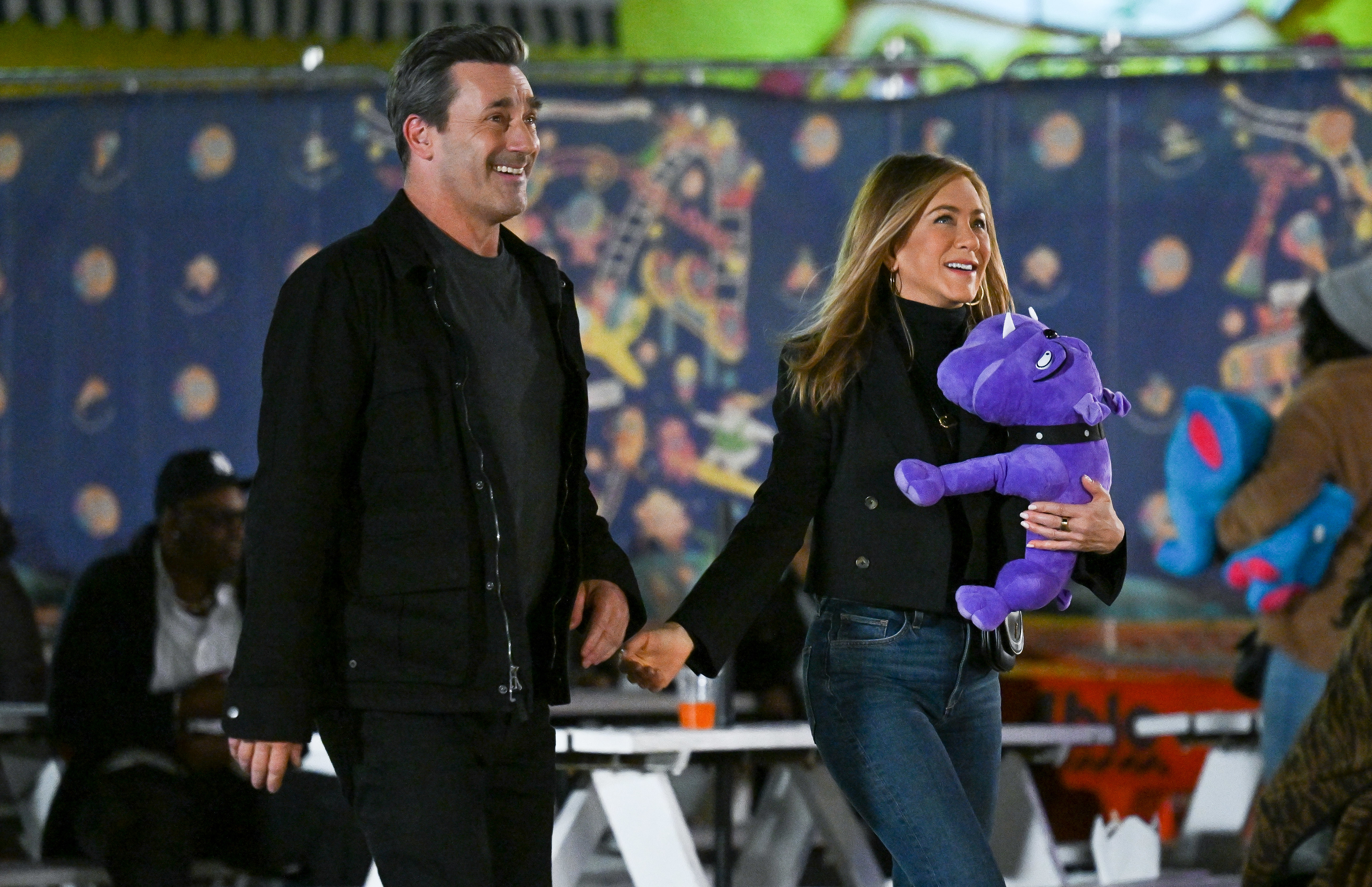 Jon Hamm and Jennifer Aniston smile while walking together at an outdoor fair. Jennifer holds a stuffed animal