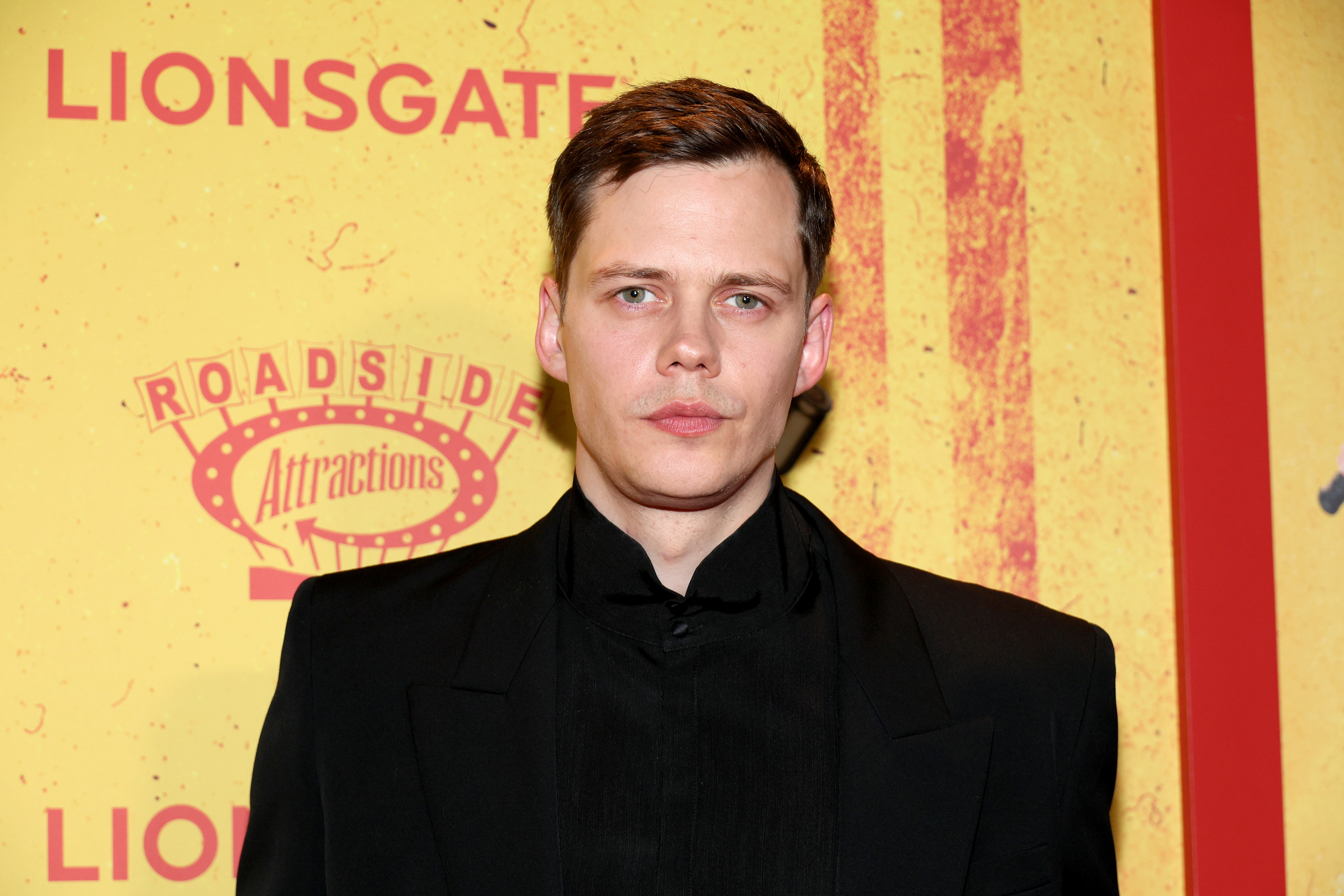 Bill Skarsgård in a black suit poses in front of a yellow backdrop featuring the Roadside Attractions and Lionsgate logos