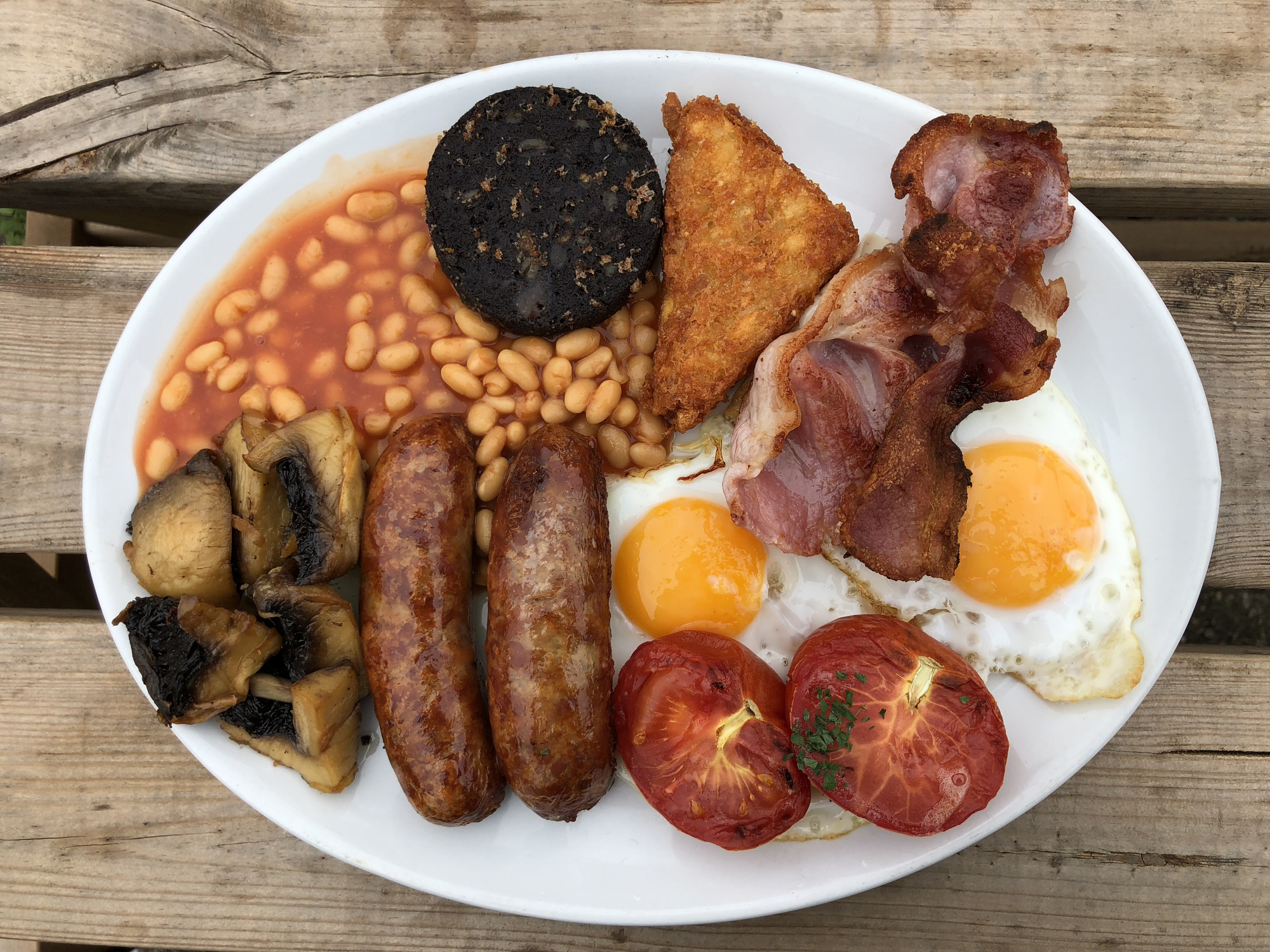 A plate filled with a traditional English breakfast: fried eggs, bacon, sausages, baked beans, grilled tomatoes, black pudding, mushrooms, and a hash brown